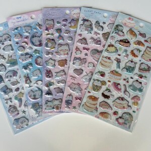 May include: Five sticker sheets with various designs. The stickers feature cute cartoon characters, food items, and celestial elements. The color palette includes pastel shades of blue, pink, purple, and white. The text "Sweet Cafe" is visible on one sheet.