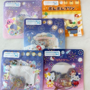 May include: A set of ten colorful, clear plastic bags filled with various character stickers. The bags are decorated with a blue and white background with fireworks and Japanese text. The characters include Cinnamoroll, Pompompurin, Little Twin Stars, and Hello Kitty.