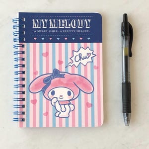May include: A pink and blue spiral bound notebook with a My Melody character on the cover. The cover has a pink and blue striped background with white hearts. The text "My Melody" is in a white banner with a blue border. The text "A sweet smile, a pretty melody." is below the banner. The character is holding a speech bubble that says "Chu!" with a heart.