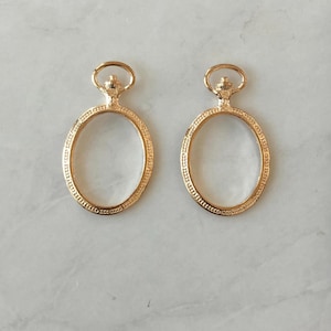 May include: Two gold-tone oval-shaped pendant frames with a decorative design around the edge. Each frame has a small loop at the top.