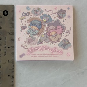 May include: A pink notepad with a Little Twin Stars illustration. The illustration features two characters with stars and clouds on a pink background. The text "Little Twin Stars" is written in a cursive font. The text below the illustration reads "When a fun day of birthday celebrations, the party continues on in their dreams..."