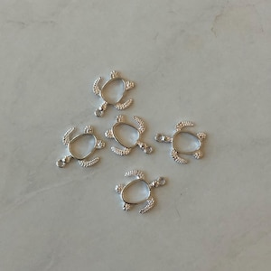 May include: Five silver turtle charms with a round opening for adding a stone or other embellishment. The charms have a loop at the top for attaching to a necklace or bracelet.