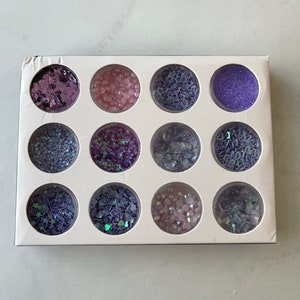 May include: A white rectangular box containing twelve circular compartments filled with various shades of purple and iridescent glitter, sequins, and beads. The box is designed for nail art or craft projects.