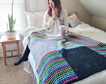 Può includere: Una donna è seduta su un letto con una coperta all'uncinetto a righe colorate. La coperta ha un quadrato nero al centro. Il letto è bianco e ha un cuscino bianco e un cuscino con un motivo.