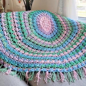 May include: A crocheted throw blanket in shades of green, pink, and blue with a fringe border. The blanket is made in a circular pattern with a central medallion.