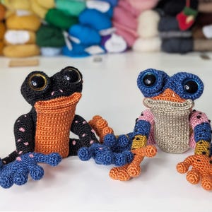 Puede incluir: Dos peluches de rana de crochet, uno negro con manchas rosas y detalles naranjas, el otro beige con detalles azules, rosas y amarillos. Ambos tienen grandes ojos azules.