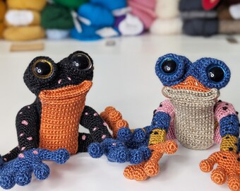 May include: Two crocheted frog plushies, one black with pink spots and orange accents, the other beige with blue, pink, and yellow accents. Both have large blue eyes.