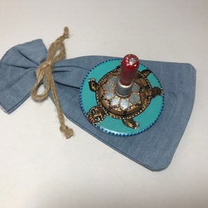 May include: A hand-painted wooden spinning top with a turtle design. The top is painted in shades of brown, gold, and blue. The turtle is on a turquoise background. The top has a red wooden handle.
