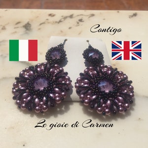 Może przedstawiać: Para kolczyków wiszących z motywem fioletowego kwiatu. Kolczyki wykonane są z małych fioletowych koralików i większego fioletowego kryształu w środku. Kolczyki wiszą na srebrnym haczyku.