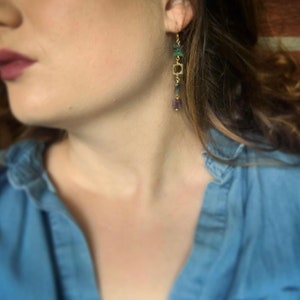 May include: A pair of gold dangle earrings with green and purple gemstone beads. The earrings feature a square charm and are hanging from the earlobe.