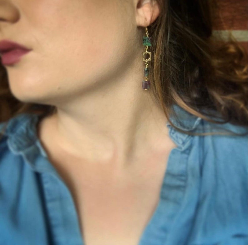 May include: A pair of gold dangle earrings with green and purple gemstone beads. The earrings feature a square charm and are hanging from the earlobe.