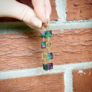 May include: Gold earrings with green and red gemstone squares. The earrings are hanging from a gold chain.