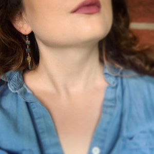 May include: A woman wearing a blue denim shirt with long, dangling earrings featuring green and purple stones.