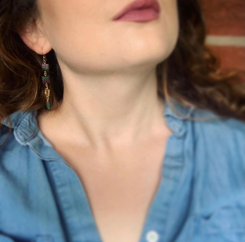May include: A woman wearing a blue denim shirt with long, dangling earrings featuring green and purple stones.