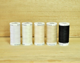Op de afbeelding: Zes spoelen Gutermann garen in wit, crème, beige en zwart. De spoelen zijn gelabeld met de merknaam en de lengte van het garen in meters en yards.