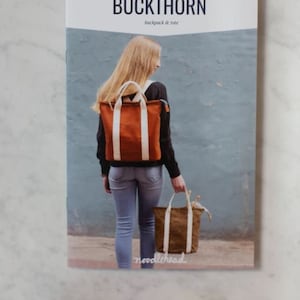 Może przedstawiać: Broszura z wzorami szycia do plecaka i torby. Na okładce znajduje się kobieta w brązowym plecaku i z brązową torbą. Tytuł wzoru to "BUCKTHORN backpack & tote", a projektant to "noodlehead".