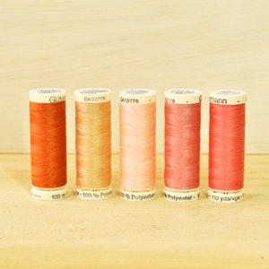 May include: Six spools of thread in various shades of orange, pink, and red. The spools are labeled with the brand name "Gutermann" and the text "100% Polyester".