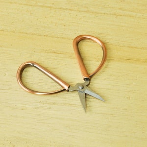 May include: A pair of small, copper-colored scissors with silver blades. The scissors are on a light wood surface.