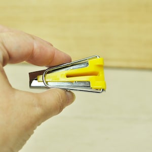 May include: A yellow and silver bias tape maker held in a hand. The tool has a yellow plastic guide and a silver metal frame. The tool is designed to fold fabric strips into bias tape.