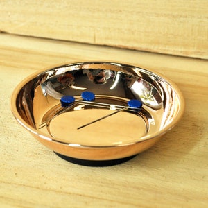 Könnte beinhalten: Eine kupferfarbene magnetische Teileablage mit schwarzem Sockel. Das Tablett enthält drei blaue Objekte und zwei Metallstäbe. Die reflektierende Oberfläche des Tabletts zeigt eine verzerrte Reflexion.