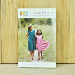 May include: A sewing pattern for a Geranium dress for children, sizes 6-12. The pattern is by Rae Hoekstra and is labelled as intermediate difficulty. The image shows two girls wearing the dress in a green and pink colour.