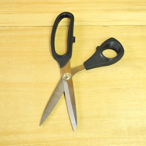 Może przedstawiać: Otwarty nożyce z czarnymi uchwytami i srebrnymi ostrzami, leżące na jasnobrązowej drewnianej powierzchni. Ostrza są ostre i gotowe do cięcia. Nożyce mają około 20 cm długości.