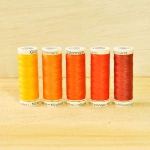 May include: Five spools of Gutermann thread in shades of yellow, orange, and red. Each spool is labeled with the brand name and the length of the thread, approximately 110 yards. The spools are arranged in a row on a light wood surface.