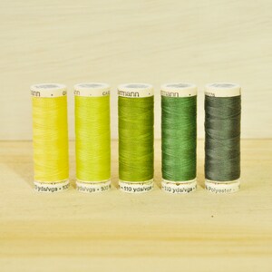Pode incluir: Seis carretéis de fio em tons de amarelo e verde. Os carretéis são etiquetados com "110 yds/vgs" e "Polyester".