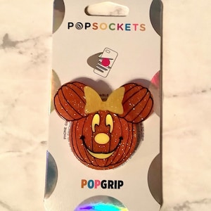 May include: A PopSockets PopGrip featuring a pumpkin-shaped Minnie Mouse design with a yellow bow. The orange and glittery grip is attached to a white backing with the PopSockets and PopGrip logos.