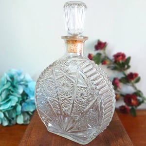 May include: A clear, round glass bottle with a decorative stopper. The bottle has a detailed, geometric pattern and a cork stopper. The stopper is clear glass. The bottle is displayed on a wooden surface.