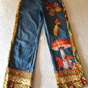 May include: Blue denim jeans featuring decorative fabric side panels with a yellow, green, and orange floral pattern. The front of the jeans is embellished with a large red and white mushroom graphic, a butterfly patch, and a small cat-like figure.
