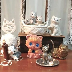 May include: A collection of ceramic cat figurines, including a pink cat with blue eyes, a white cat with a basket, and a gray cat with a brown bottle. The figurines are arranged on a wooden surface.