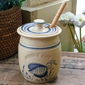 May include: A vintage honey pot with a lid and a wooden honey dipper. The stoneware pot is beige with blue stripes and a blue bee illustration. The words "HONEY POT" are written on the front.