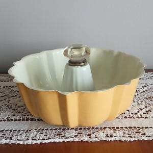 May include: A yellow fluted baking mold with a central tube and a clear glass knob handle on top. The interior of the mold is a pale cream color, and the exterior features a smooth, solid yellow finish with a scalloped, wavy rim design.