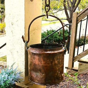 May include: A cylindrical copper pot with a hammered texture and a dark, weathered patina. The pot features a sturdy, arched metal handle attached to two side loops, suspended from a metal hook by a series of connected iron rings.