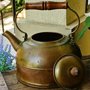 May include: Antique brass kettle with a wooden handle and lid. The kettle has a rounded body, a spout, and a curved handle. The lid is off to the side. The kettle is sitting on a wooden surface.