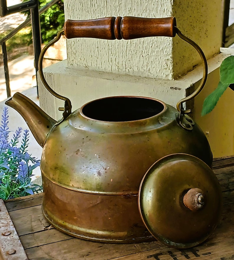 May include: Antique brass kettle with a wooden handle and lid. The kettle has a rounded body, a spout, and a curved handle. The lid is off to the side. The kettle is sitting on a wooden surface.