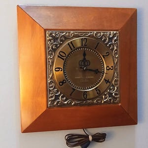 May include: A square wall clock with a smooth, light brown wooden frame and a central brass-colored face. The face features black Arabic numerals, ornate silver-toned corner detailing, and a brown electrical cord hanging from the bottom.