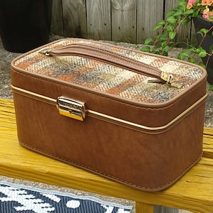 May include: A vintage brown makeup case with a plaid top and gold-tone hardware. The rectangular case has a handle and a clasp closure. The case is sitting on a yellow bench.