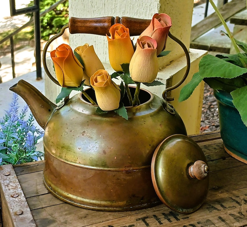 May include: An antique brass kettle with a wooden handle and lid, filled with wooden roses in shades of orange, yellow, and peach. The kettle sits on a wooden surface, with a green plant in a pot visible in the background.