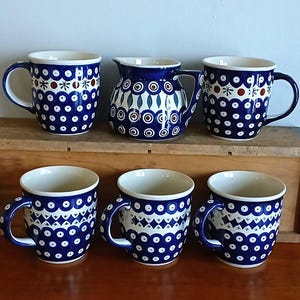 May include: A set of five ceramic mugs and one small pitcher featuring a dark blue and white pattern with repeating circles, dots, and geometric shapes. The interior of each piece is cream-colored, and each mug has a single handle on the side.