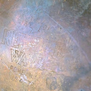 May include: Close-up of a tarnished metal surface with the embossed text "REVERE WARE" and "ROME, N.Y. USA-78". The surface has a mottled appearance with shades of brown, blue, and green. The numbers "1801" are also visible.