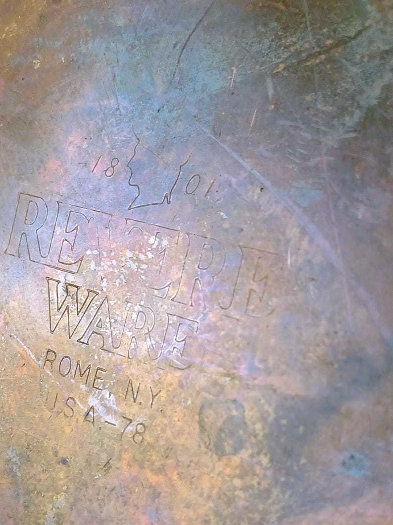 May include: Close-up of a tarnished metal surface with the embossed text "REVERE WARE" and "ROME, N.Y. USA-78". The surface has a mottled appearance with shades of brown, blue, and green. The numbers "1801" are also visible.