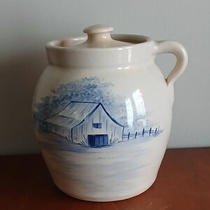 May include: A cream-colored ceramic crock with a matching lid and a single side handle. The exterior features a hand-painted blue illustration of a barn surrounded by trees and a fence, rendered in a soft, watercolor-style brushwork technique.