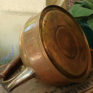 May include: A vintage copper kettle with a wooden handle and spout. The kettle has a rounded body and a shiny, aged finish. The spout is made of a lighter-colored metal. The kettle is resting on a wooden surface.