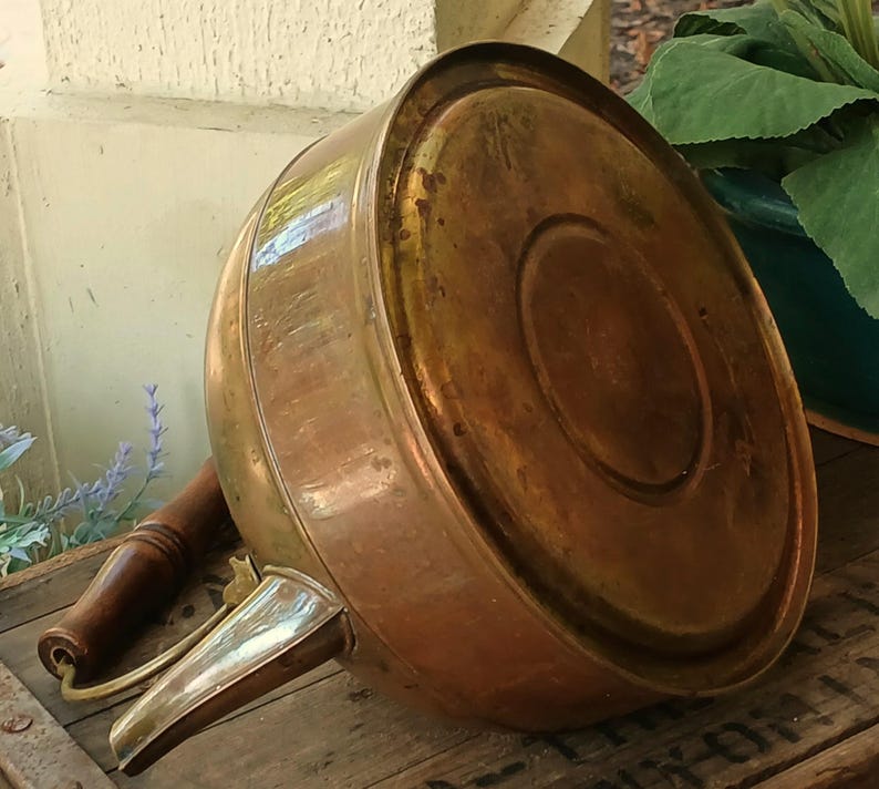 May include: A vintage copper kettle with a wooden handle and spout. The kettle has a rounded body and a shiny, aged finish. The spout is made of a lighter-colored metal. The kettle is resting on a wooden surface.