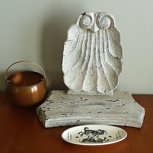 May include: A white distressed wooden owl-shaped display stand with a scalloped texture, a small copper-colored metal bowl with a handle, and an oval white ceramic dish featuring a black graphic print of two goats and the text WOULD YOU LIKE TO