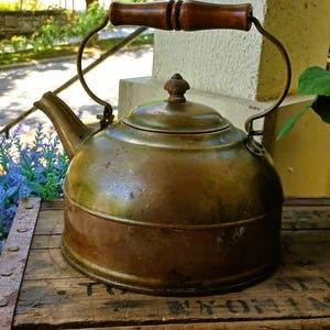 May include: Antique copper kettle with a wooden handle and lid. The kettle has a curved spout and a metal handle. The copper has a patinaed finish, giving it a vintage look. The kettle sits on a wooden crate.