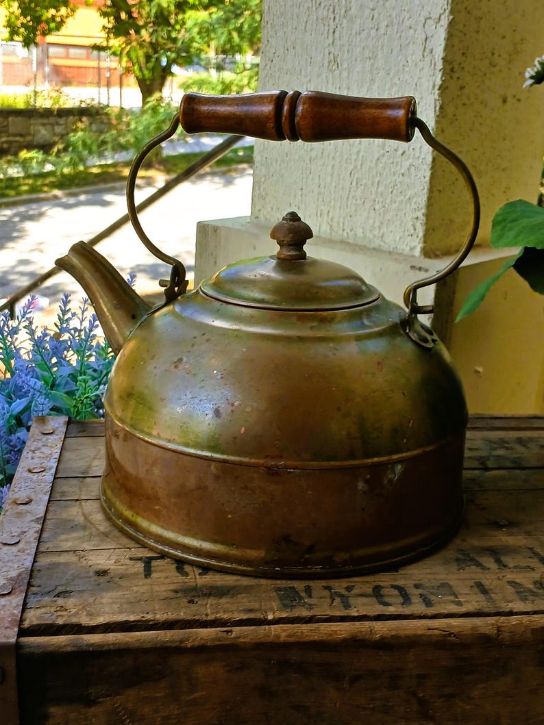 May include: Antique copper kettle with a wooden handle and lid. The kettle has a curved spout and a metal handle. The copper has a patinaed finish, giving it a vintage look. The kettle sits on a wooden crate.