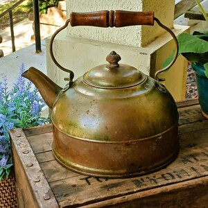 May include: Antique brass kettle with a wooden handle. The kettle has a rounded body, a spout, and a lid with a small knob. The handle is made of two pieces of wood joined together. The kettle sits on a wooden crate.